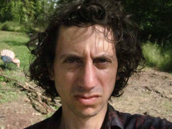 A close up of an adult man who isn't smiling, poses for a photo. His hair is dark, short, and curly.