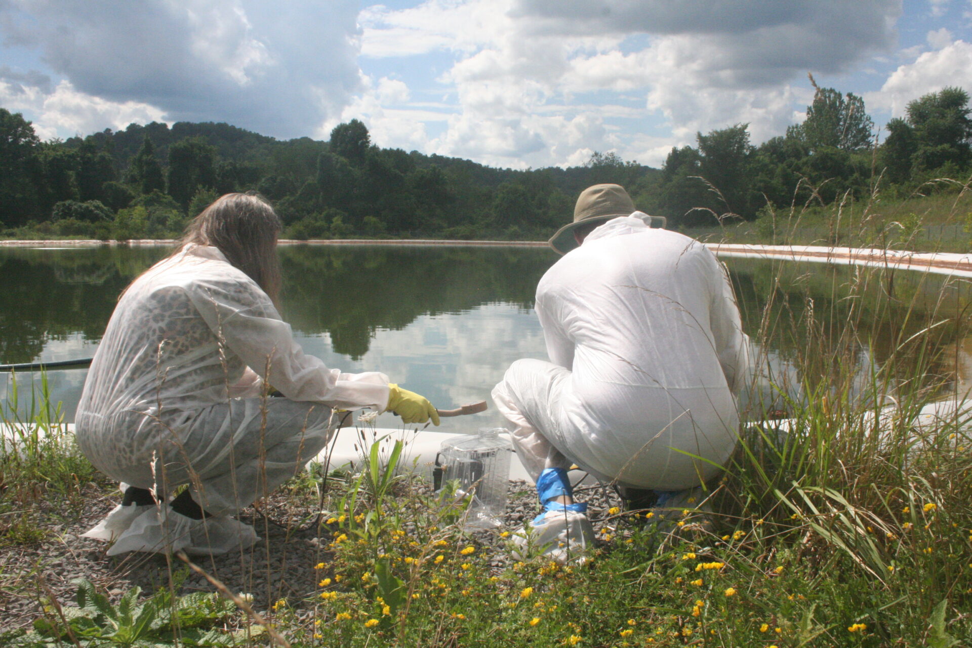 Two adults, with their backs to the camera, wear protective suits and coverings while leaning in to collect samples in water.