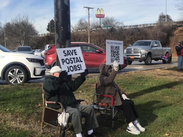 Two individuals sit in lawn chairs on the side of the road, holding up signs to spread awareness about the proposed downsizing of a Charleston mail processing facility.
