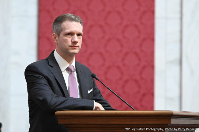 A man wearing a dark gray suit and light purple tie stands at a podium with a marbled wall covered by red tapestry in the background