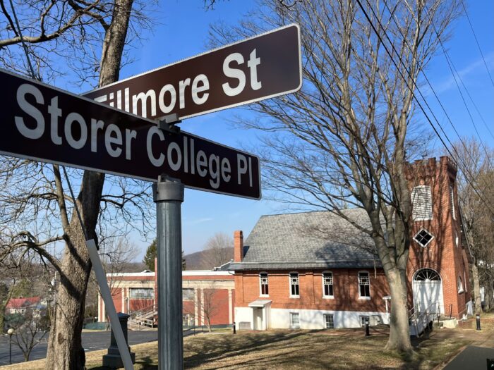 A road sign for Storer College Place sits in the foreground. Behind it stands the Curtis Freewill Baptist Church, with a few trees in between.