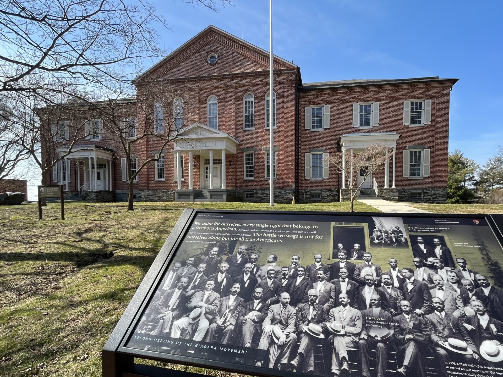 A sign displaying a photograph of participants in the second meeting of the Niagara Movement stands outside of Anthony Hall, a brick building on the campus of Storer College.