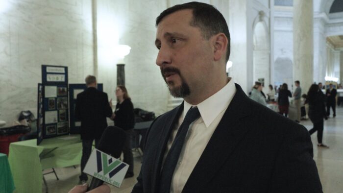 A man with a goatee, wearing a jacket and tie, speaks in a large space with marble walls and dim light.