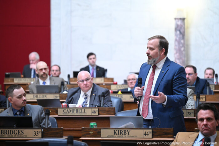 Man in suit with a red tie and wispy beard standing and speaking in large room