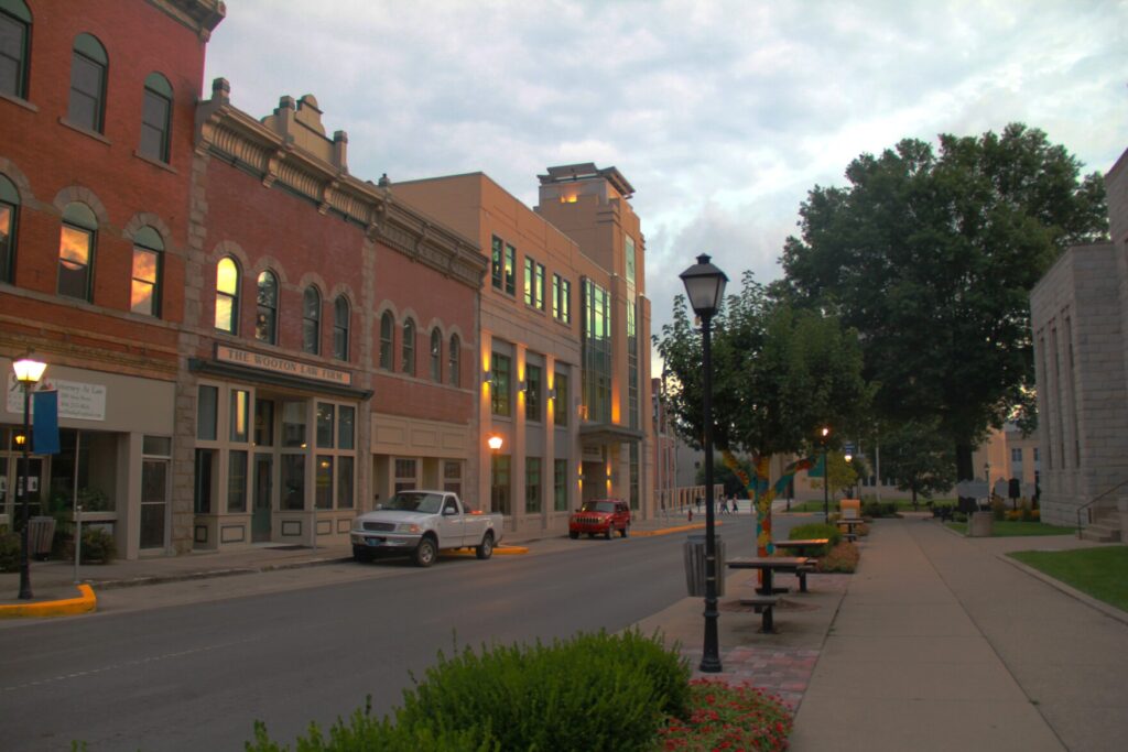 A rendering of a city street in Beckley. Buildings span the left side of the image. On the opposite side, divided by a road, there is a sidewalk with lamps, tables and trees beside it.