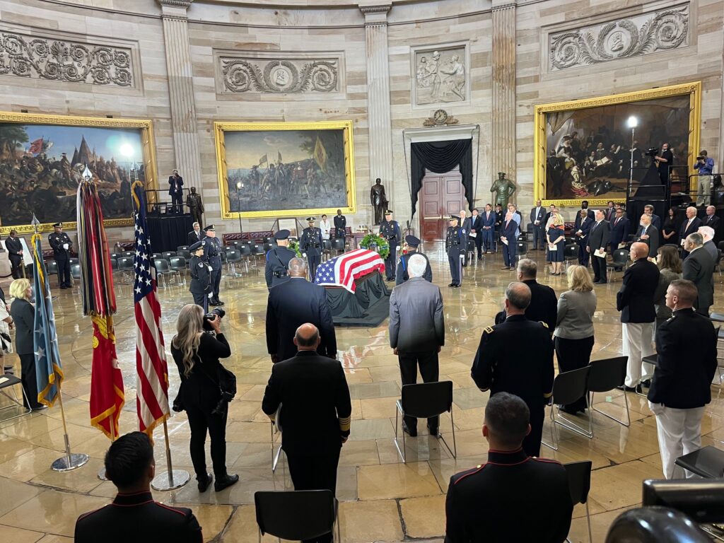 A flag-draped casket lies in the center of a round marbled space, surrounded by Marines, elected leaders, guests, reporters and photographers.
