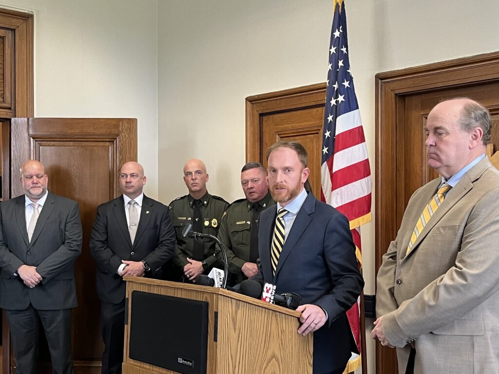 Dressed in a suit, State Auditor JB McCuskey stands at a podium, addressing members of the press of screen. He stands beside representatives from the United States Attorney's Office, Truist Bank and the West Virginia State Police, who are wearing suits and uniforms, respectively.