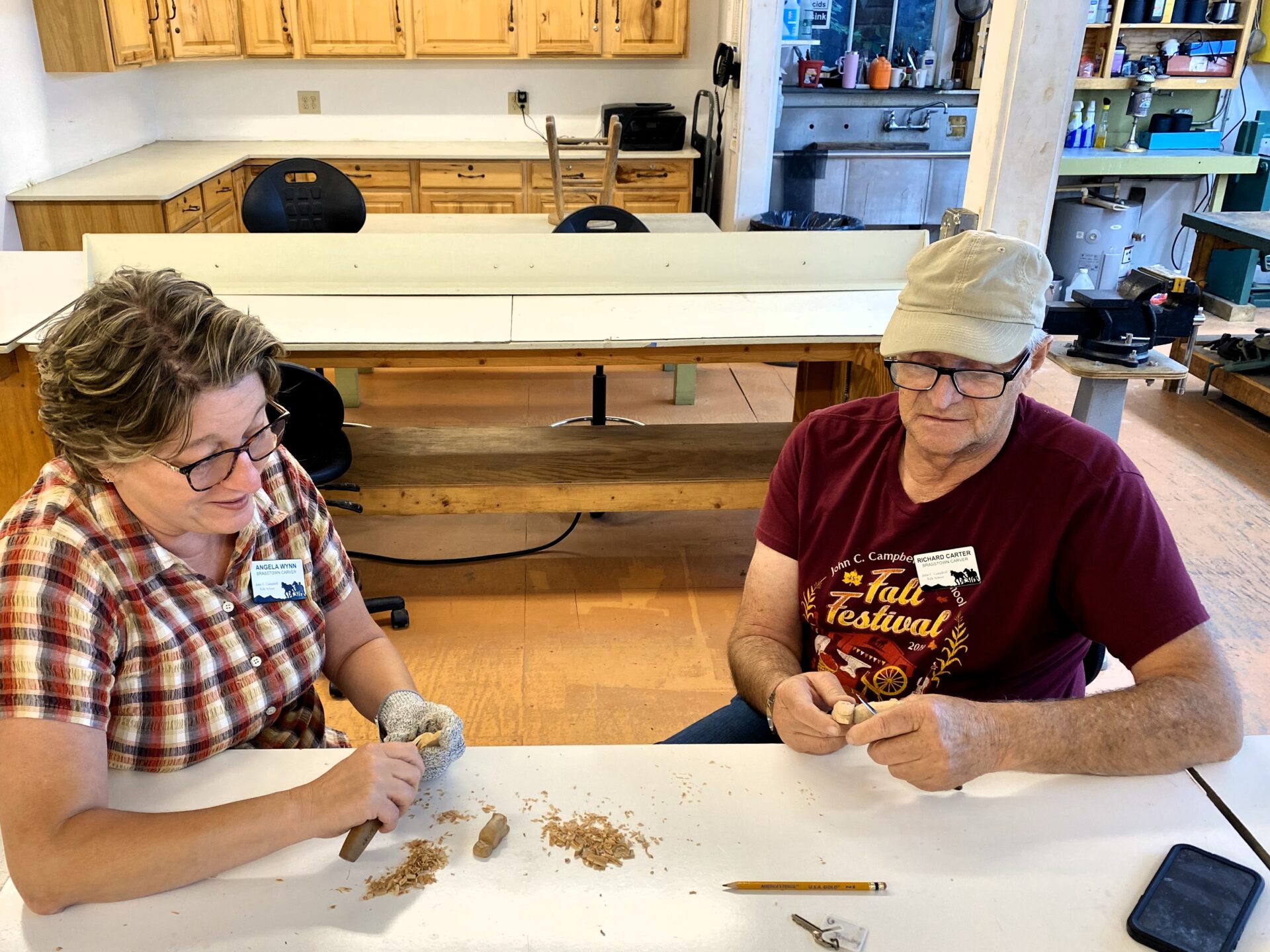 Two older adults sit at a table carving wood. One adult is female and the other is male.