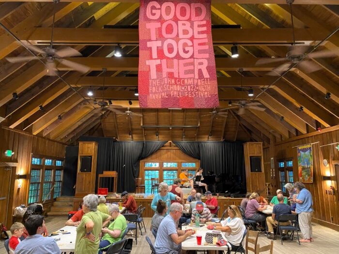 A gathering room. Dozens of people sit at chairs and tables crafting. A large sign hangs from the ceiling that reads, "Good to be together."