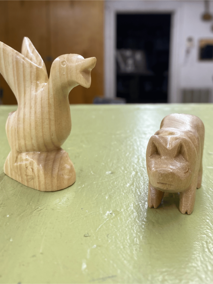 Two wood carvings are shown on a table. One is a goose while the other is a pig.