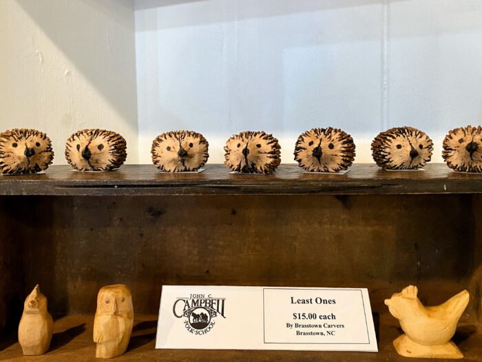 Seven small wood carvings of hedgehogs line the top of a desk. Underneath are other small carvings and a sign that reads, "Least Ones, $15.00 each by Brasstown Carvers, Brasstown, NC."