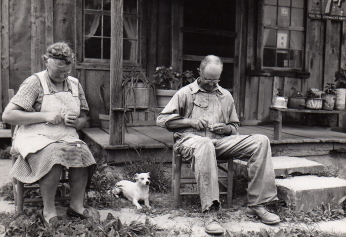 A black and white photo of an elderly man and woman sitting outside, in front of a porch carving small pieces of wood. Between them is a small white dog.