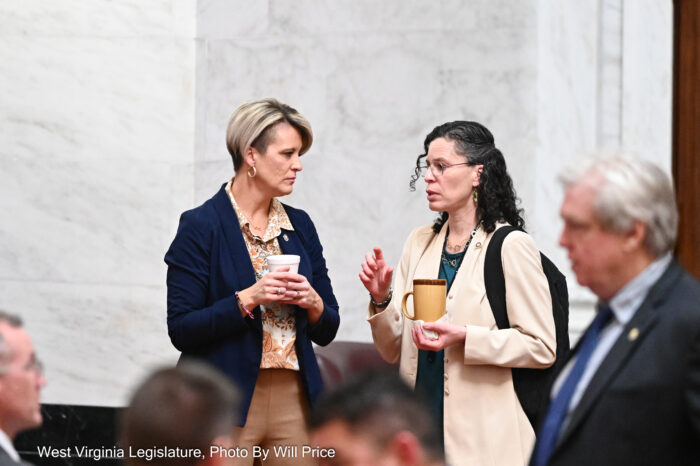 Two women stand holding coffee cups speaking to each other.