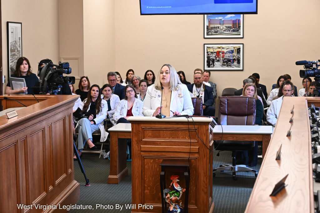 A committee room full of doctors wearing their white coats surrounds Dr. Lisa Costello as she testifies before the Senate Health Committee.