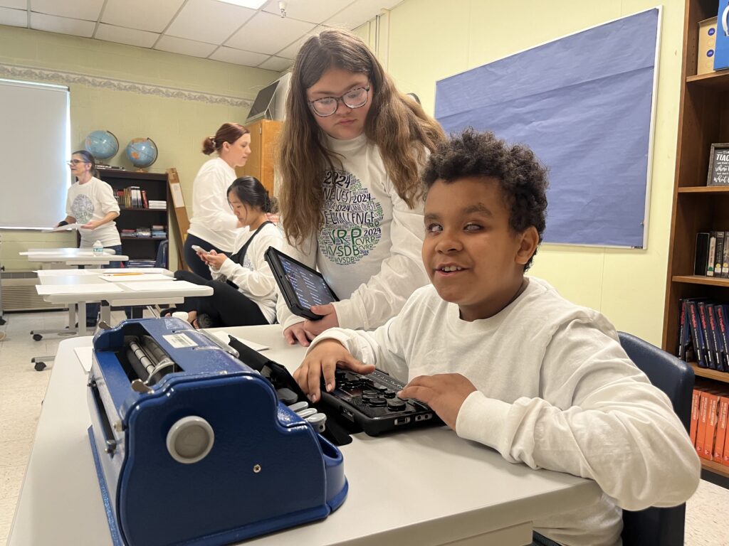A student sits at a desk and uses a Polaris machine, which is a long, flat tablet with finger pads. Another student stands behind him, watching.