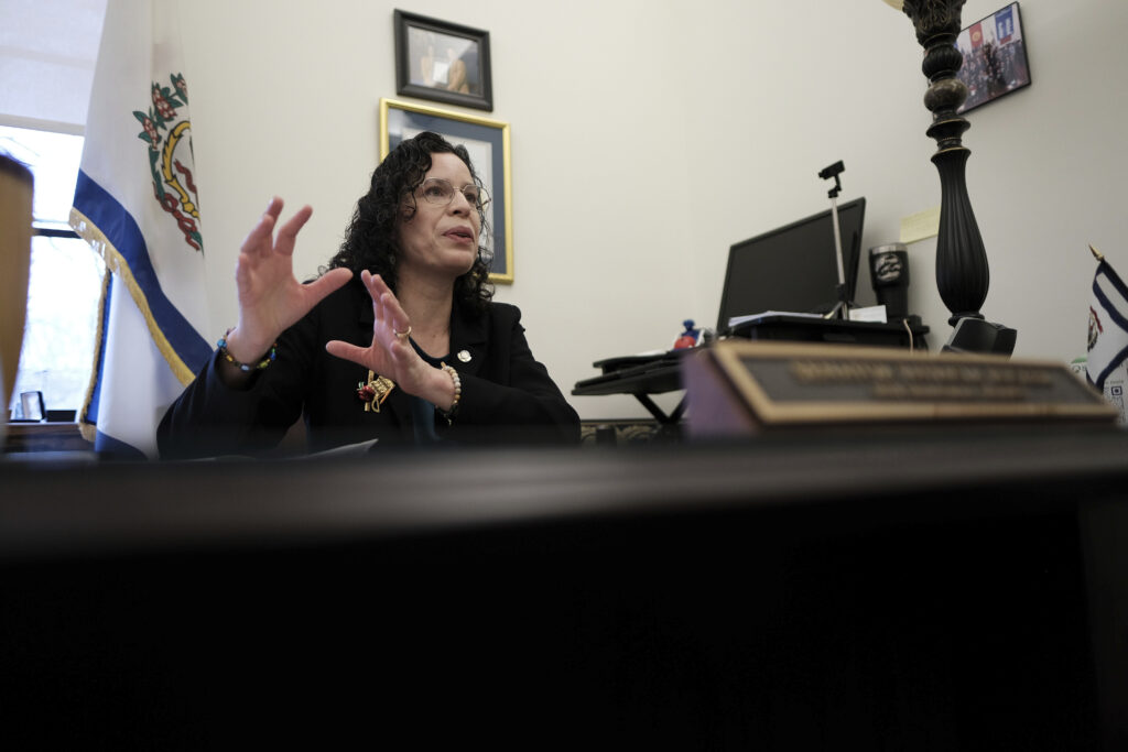 A woman with dark hair sits at her desk. She gestures with her hands as if she is speaking with someone off camera.