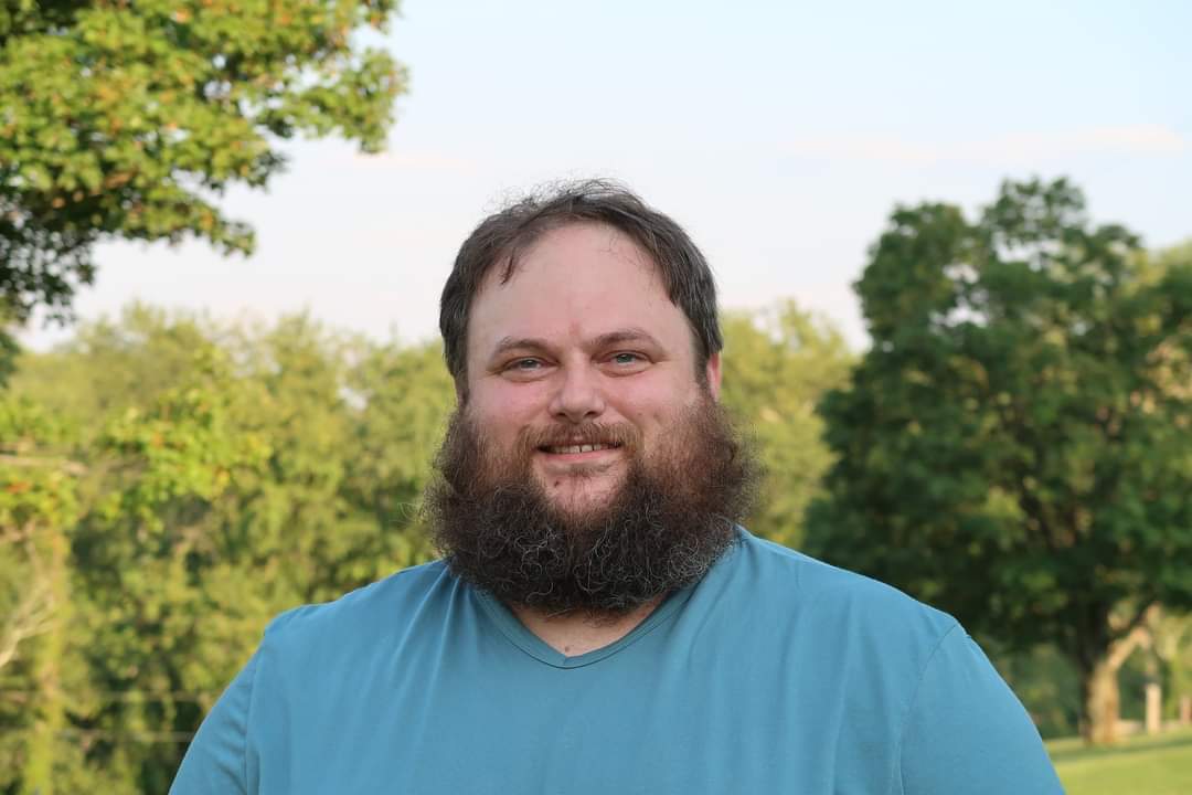 An adult person in a blue shirt poses for a photo. They have brown hair and a brown beard. Behind them is a blue sky and green trees.
