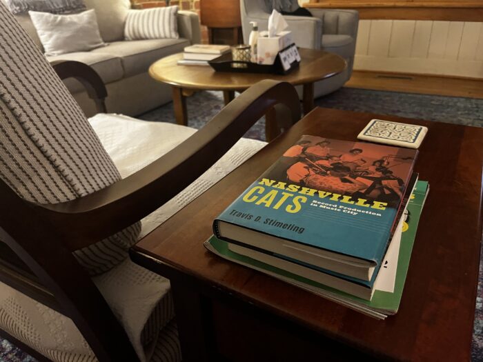 A stack of books are shown on an end table next to a chair. The book on the top of the stack reads, "Nashville Cats by Travis D. Stimeling."