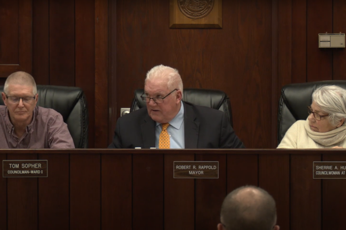 Seated beside councilmembers at a desk, Mayor Rob Rappold turns his head to address a speaker out of the camera frame.