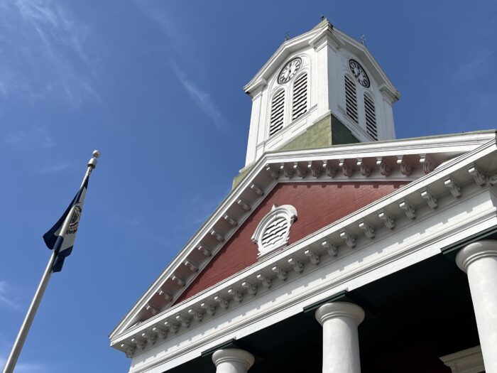 With four columns and a spire, the Jefferson County Courthouse stands before a clear blue sky. The flag of West Virginia stands to its side.