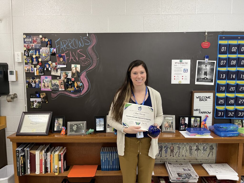 A young female teacher stands in front of a blackboard. She holds a certificate in her hand and a blue apple paperweight.