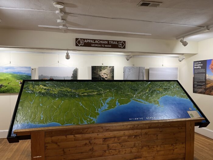 A relief map displaying the Appalachian mountain range stretches across a room with wooden floors and photographs along the walls. On the wall behind it, a sign reads "Appalachian Trail," with an arrow marked "South" pointed toward Georgia and an arrow marked "North" pointed toward Maine.
