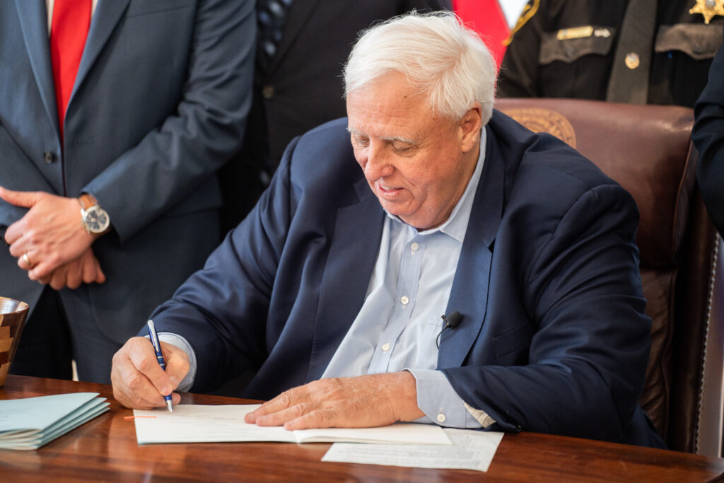 Governor Jim Justice sits at his desk. He is signing a piece of paper with a pen in his right hand.