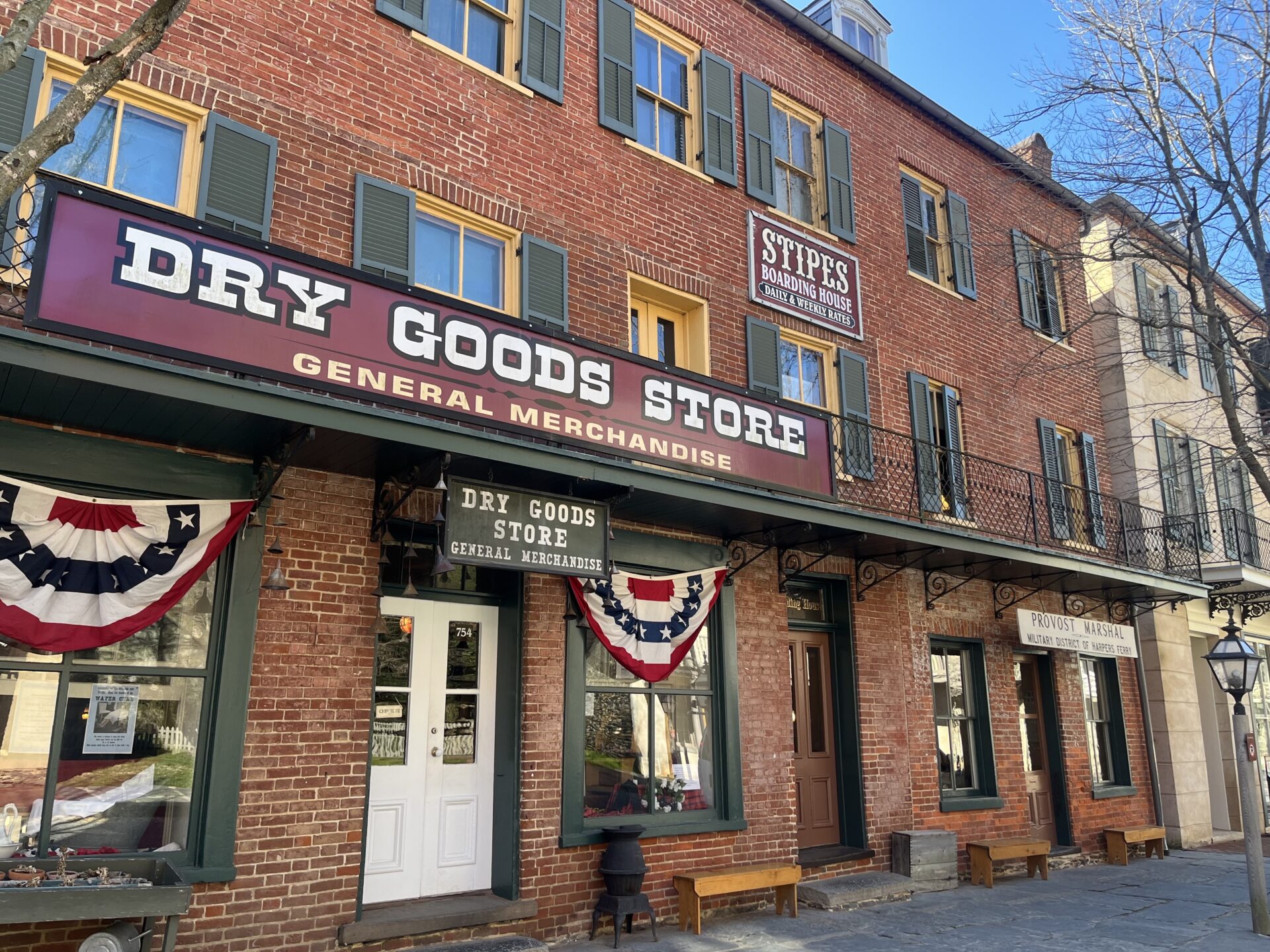 Olden brick buildings display fake, old-time signs that read "Dry Goods Store" and "Stipes Boarding House." The buildings line a street in downtown Harpers Ferry, with a stone sidewalk before them.