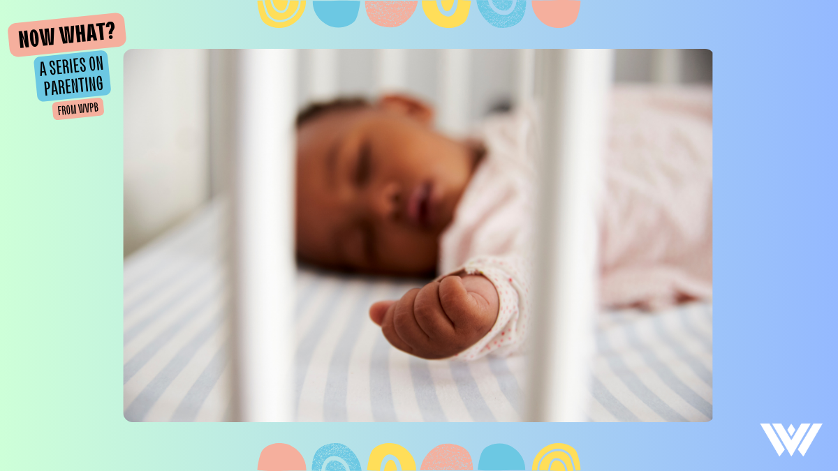 A close up of a sleeping baby in her crib. Her face is blurred slightly, while her hand is in focus. She wears a pink polka dot onesie, and her sheets feature light blue and white stripes.