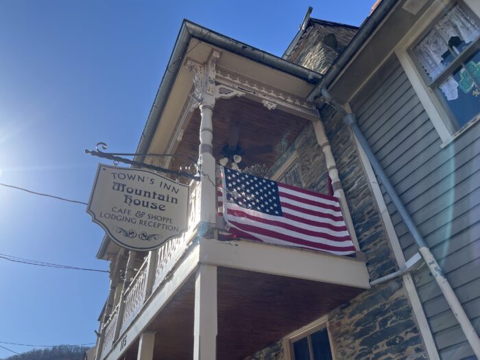 A balcony extends out from a second-floor hotel room inside a stone building. On one side of the balcony hangs an American flag. On another side is a sign that reads in an old-time font: "Town's Inn Mountain House" and "Cafe & Shoppe, Lodging, Reception."