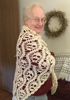 An elderly woman with white hair smiles for the camera and is wrapped in a knitted, white shawl.
