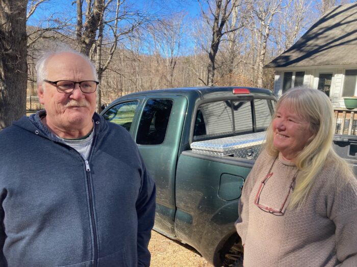 Two older people with white hair stand for a photo. They appear to be talking. Behind them is a green pickup truck. The man wears a blue hoodie and black glasses. The woman wears a pink sweater and brown glasses can be seen around her neck.
