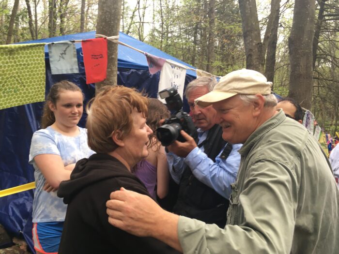 An older woman with red hair is shown beginning to hug a man with white hair and wearing a white ball cap. A photographer next to them takes their picture and a crowd of people are near them.