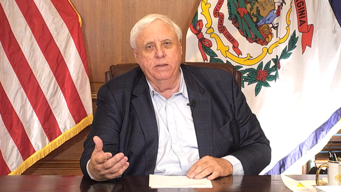 Governor Jim Justice sits at his desk, gesturing with his right hand. Behind him are the American and West Virginia flags. He is wearing a suit with the top button of his shirt unbuttoned.