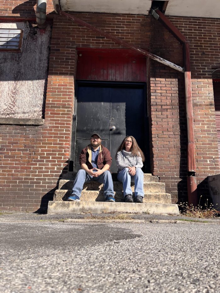 Two adults, a man and a woman, sit on the concrete steps of a brick building. They are wearing casual clothing - jeans, sweatshirts, a jacket. The man is wearing a ball cap, and the woman has long, brown hair.
