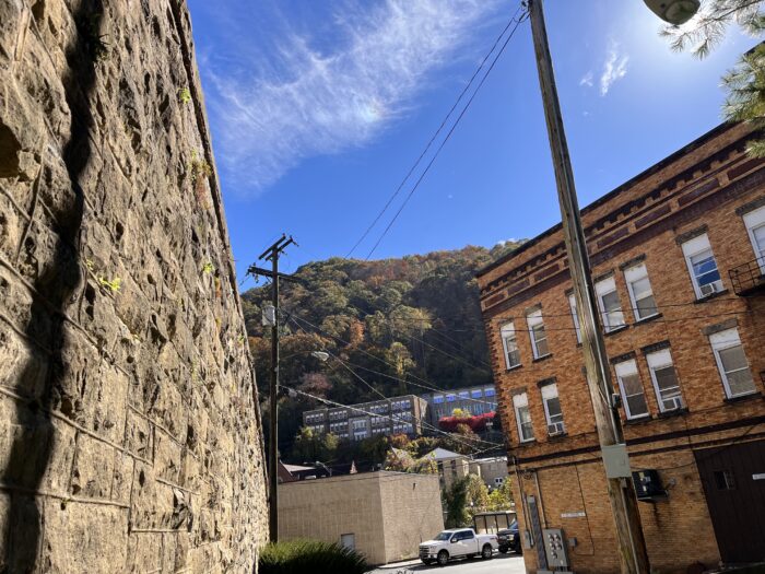 An artistic view of a mountain from downtown Welch, West Virginia. On one side of the frame is a stone wall, and in the distance on the other side is a brick building with several windows. Many of the windows have air conditioners in them.