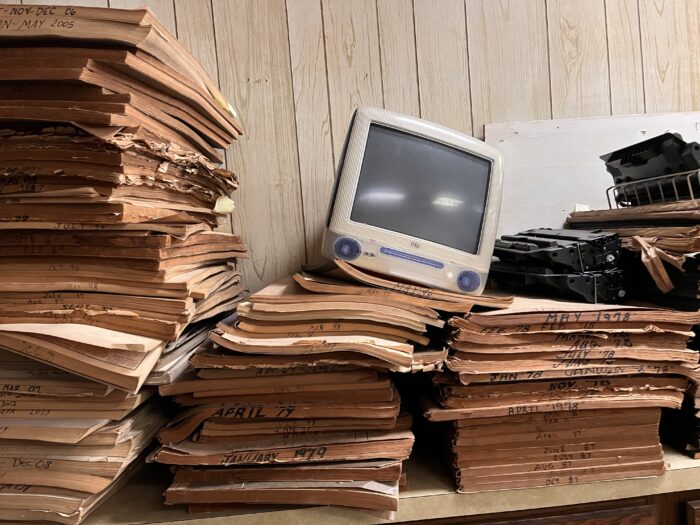 An old iMac computer is seen balanced on stacks of old paper. The iMac looks as if it is from the 1990s.