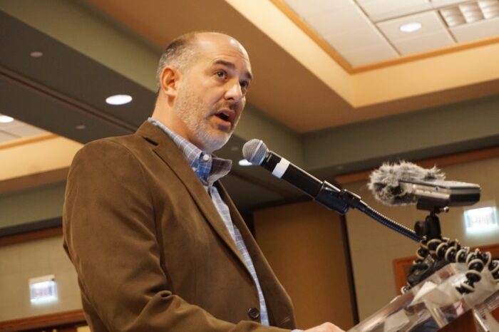 An adult man, balding, trimmed gray beard, speaks into a microphone at an event. He stands at a podium.