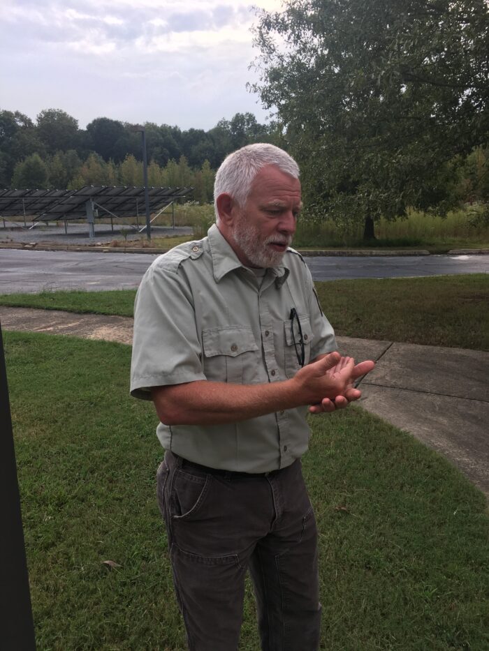 An older, adult man with white hair kept trimmed short stands on grass and gestures as if he is speaking to someone. He wears a light green button up shirt.