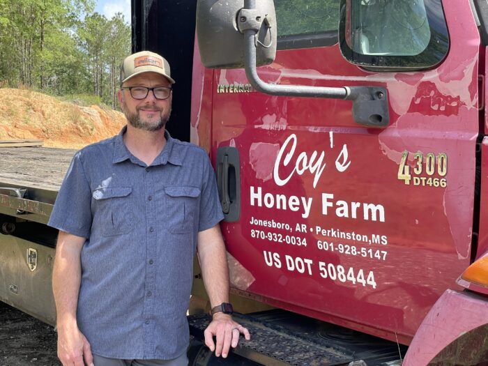 A middle age man wearing a ball cap and glasses stands next to a red semi truck. On the truck door, there is text that reads, "Coy's Honey Farm, Jonesboro, AR, Perkinston, MS, 870-932-0034, 601-928-5147."