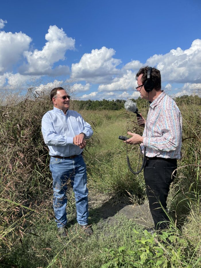 Two adult men stand next to each other in a field. The sky is blue with some white clouds. One man has headphones on and holds a recorder. The other man answers questions.