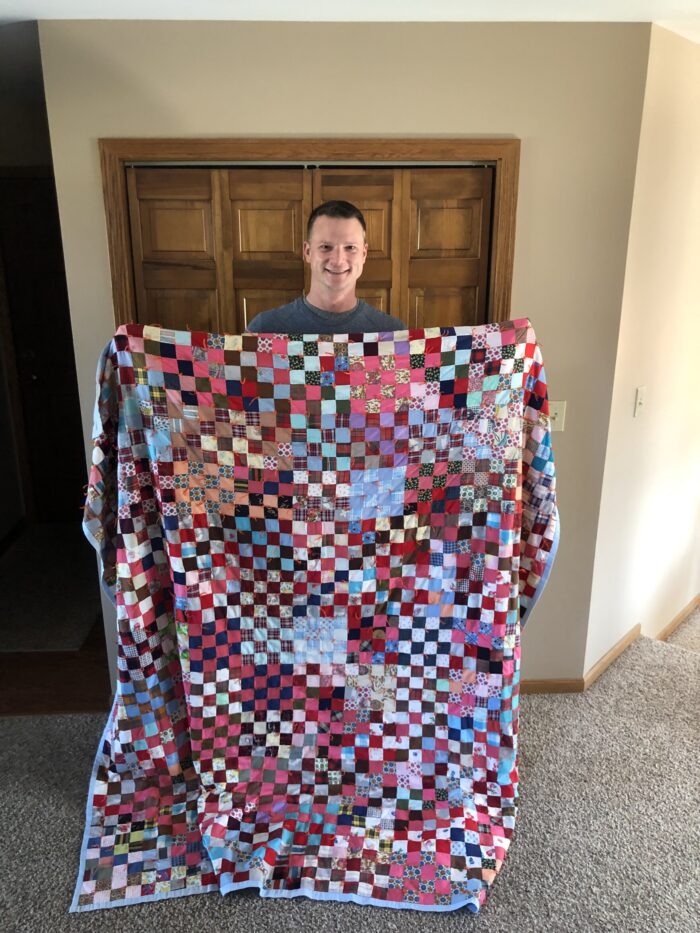 An adult man is shown smiling for the camera and holding up a quilt. The quilt shows all sorts of colors - red, blue, yellow, pink, purple, among others.
