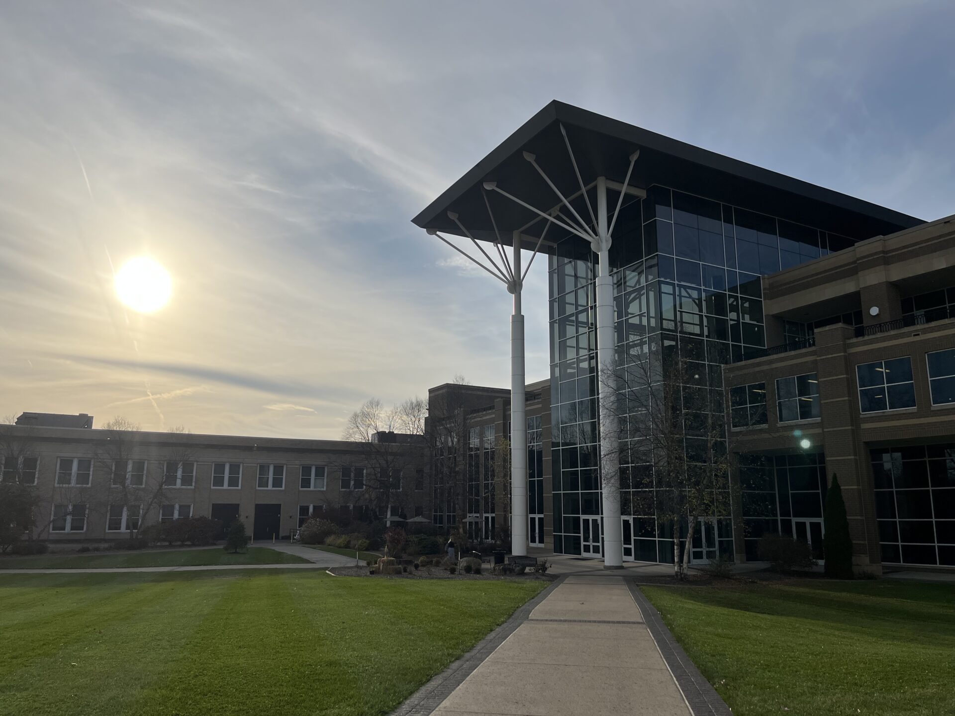 The sun sets over Fairmont State University's campus. A building with two tall columns and long, glass panes out front reflects the sunset. A cement path cuts through a grass quad, leading to the building.