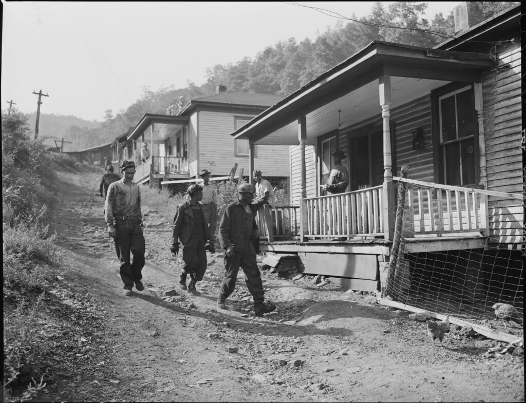 Dusted from mining coal, a group of men walk downhill on a dirt path, past houses whose residents watch them from porches.
