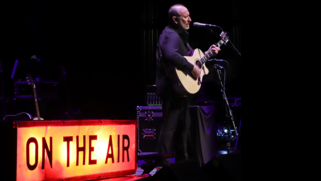 A lone man stands on a stage playing a guitar. Off the side of him is an "On The Air" sign that is lit up.