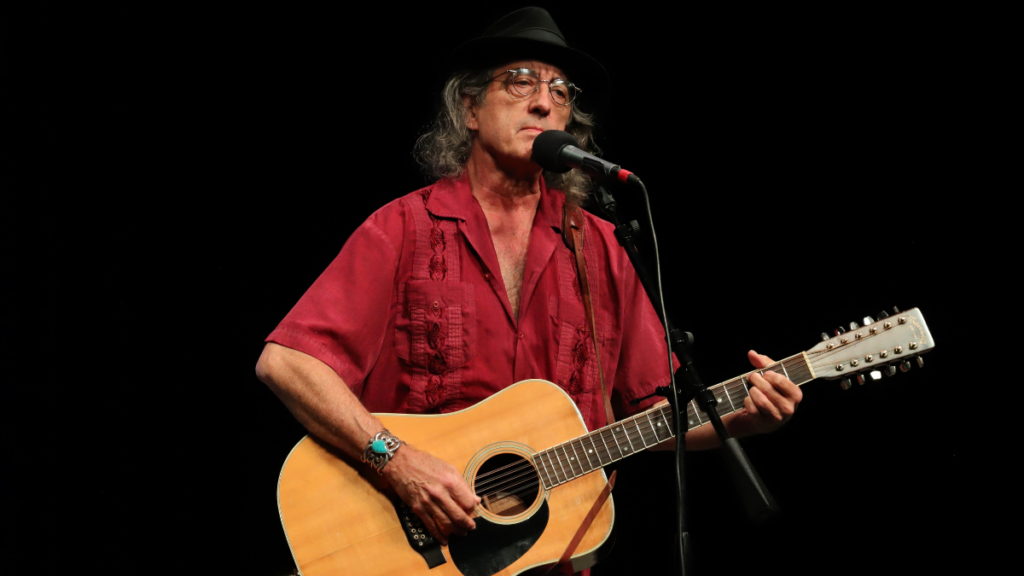 A man with a black hat, glasses, and a red button down shirt holds a guitar and sings into a mic.