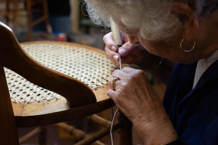 An elderly woman is leaned down close to the seat of a chair. She is weaving a new seat onto an old hand caned chair.