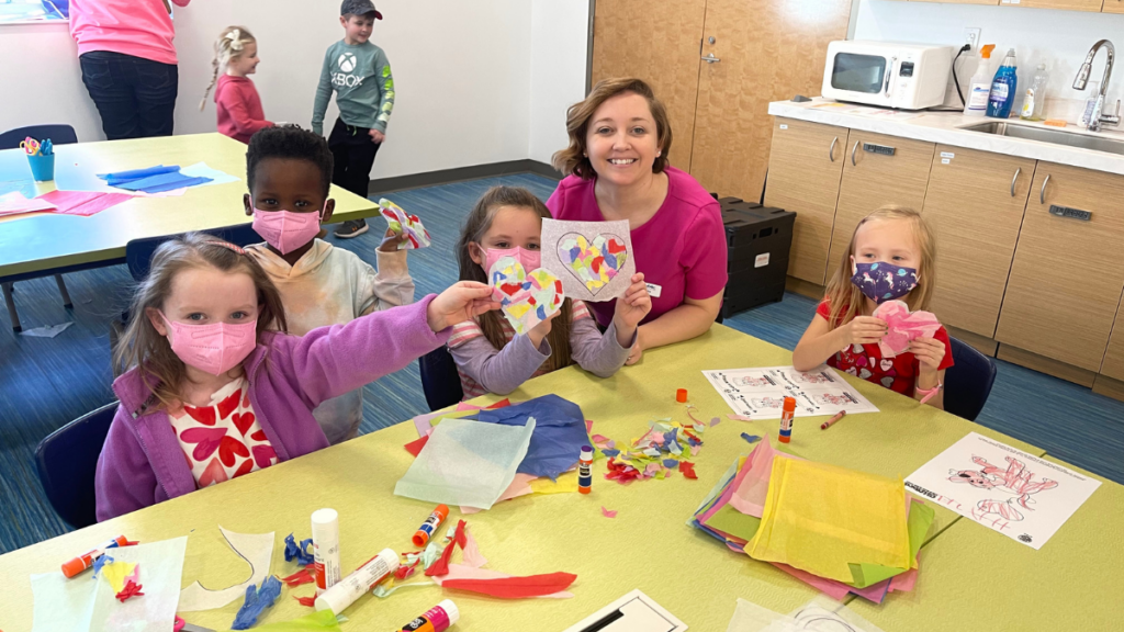 An adult woman smiles for the camera. Four young children doing crafts at a table, wearing masks, show off their artwork for the camera.