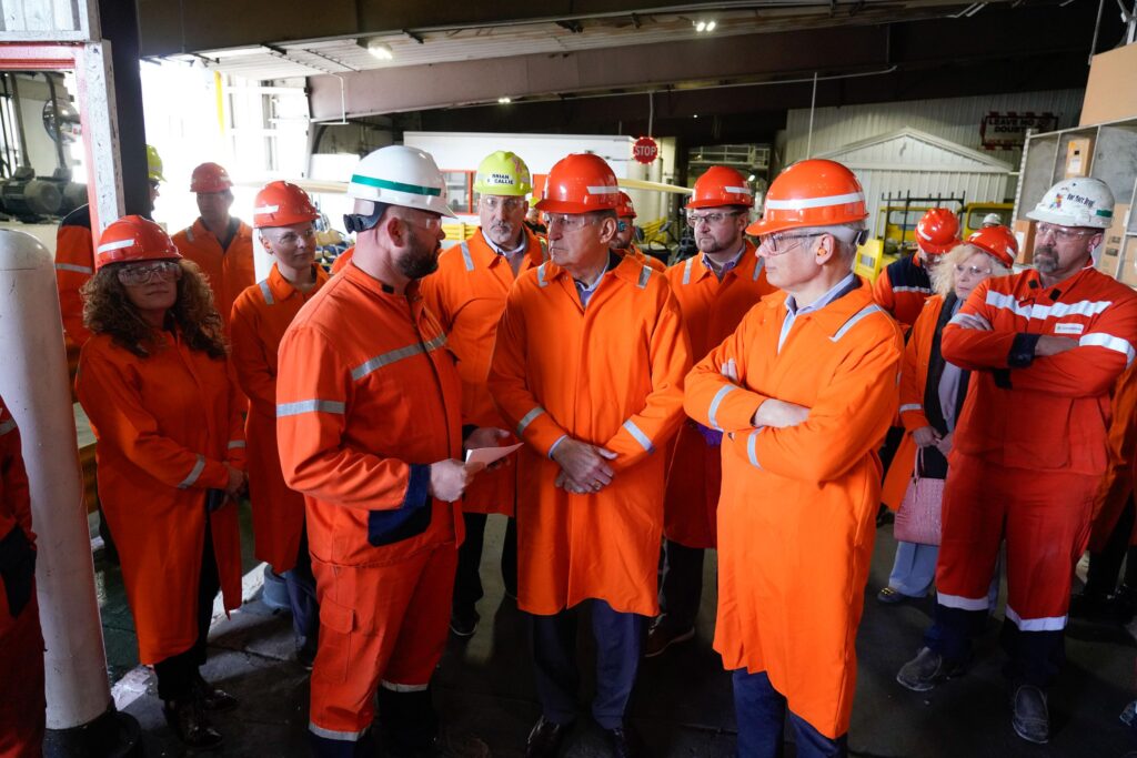 People wearing orange coats and orange hard hats gather inside an industrial building.