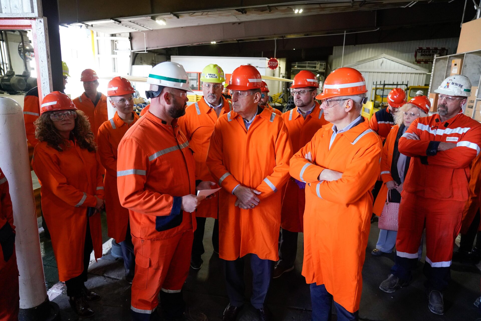 People wearing orange coats and orange hard hats gather inside an industrial building.